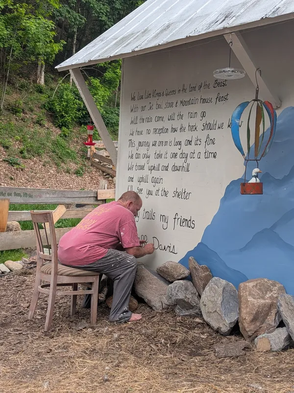 Jeff Davis sitting on a wooden chair, painting his Ode to the AT poem onto the side of the goat shed
